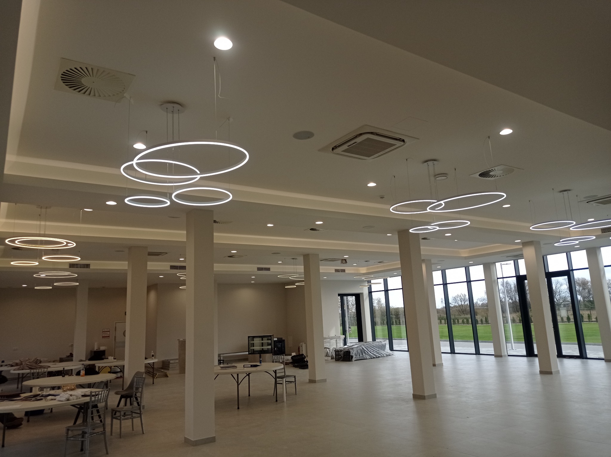 Przestronne wnętrze sali z kolumnami i nowoczesnym oświetleniem w formie okręgów. Widoczne okna z widokiem na zieleń i przygotowane stoły.