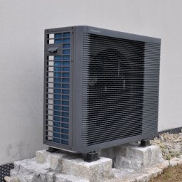 Woltbar Michał Kotewicz - Zewnętrzna jednostka pompy ciepła Panasonic Aquarea Split, zamontowana na kamiennej podstawie przy ścianie budynku. Widoczny fragment rynny i siatka.