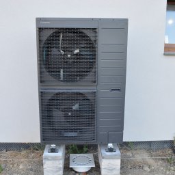 Woltbar Michał Kotewicz - Szara pompa ciepła Panasonic Aquarea Split, zamontowana na betonowych blokach przy ścianie budynku z widocznym oknem. Widoczne elementy instalacji odprowadzającej skropliny.