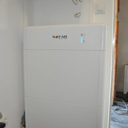 Woltbar Michał Kotewicz - Biały akumulator energii SoFar Solar z diodami LED, widoczny w pomieszczeniu w trakcie instalacji. W tle odkurzacz przemysłowy i elementy wykończeniowe.