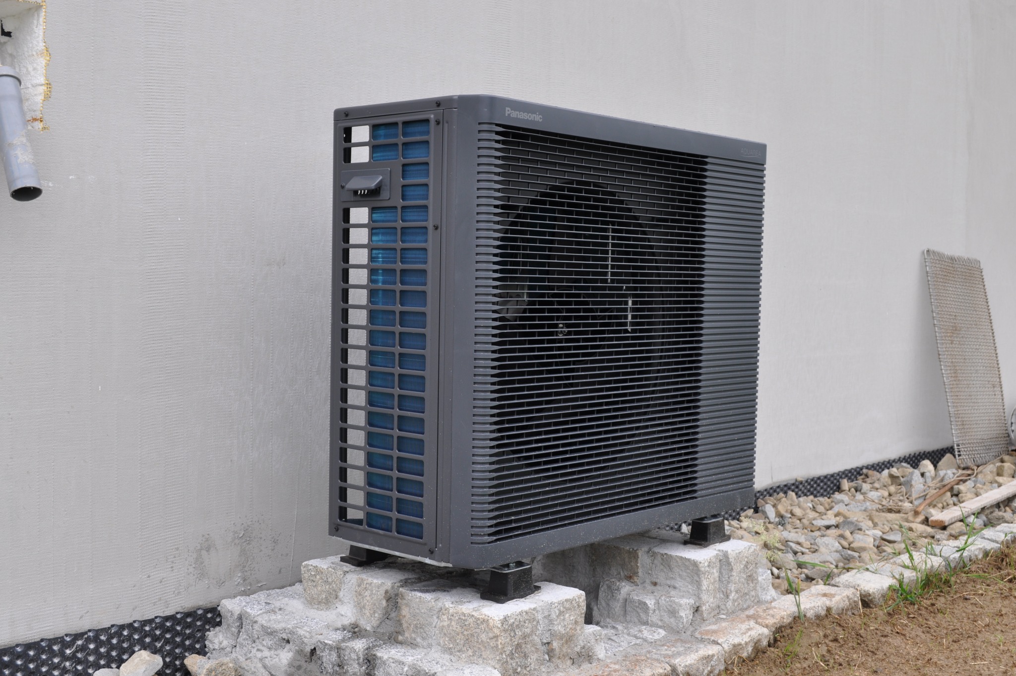 Zewnętrzna jednostka pompy ciepła Panasonic Aquarea Split, zamontowana na kamiennej podstawie przy ścianie budynku. Widoczny fragment rynny i siatka.