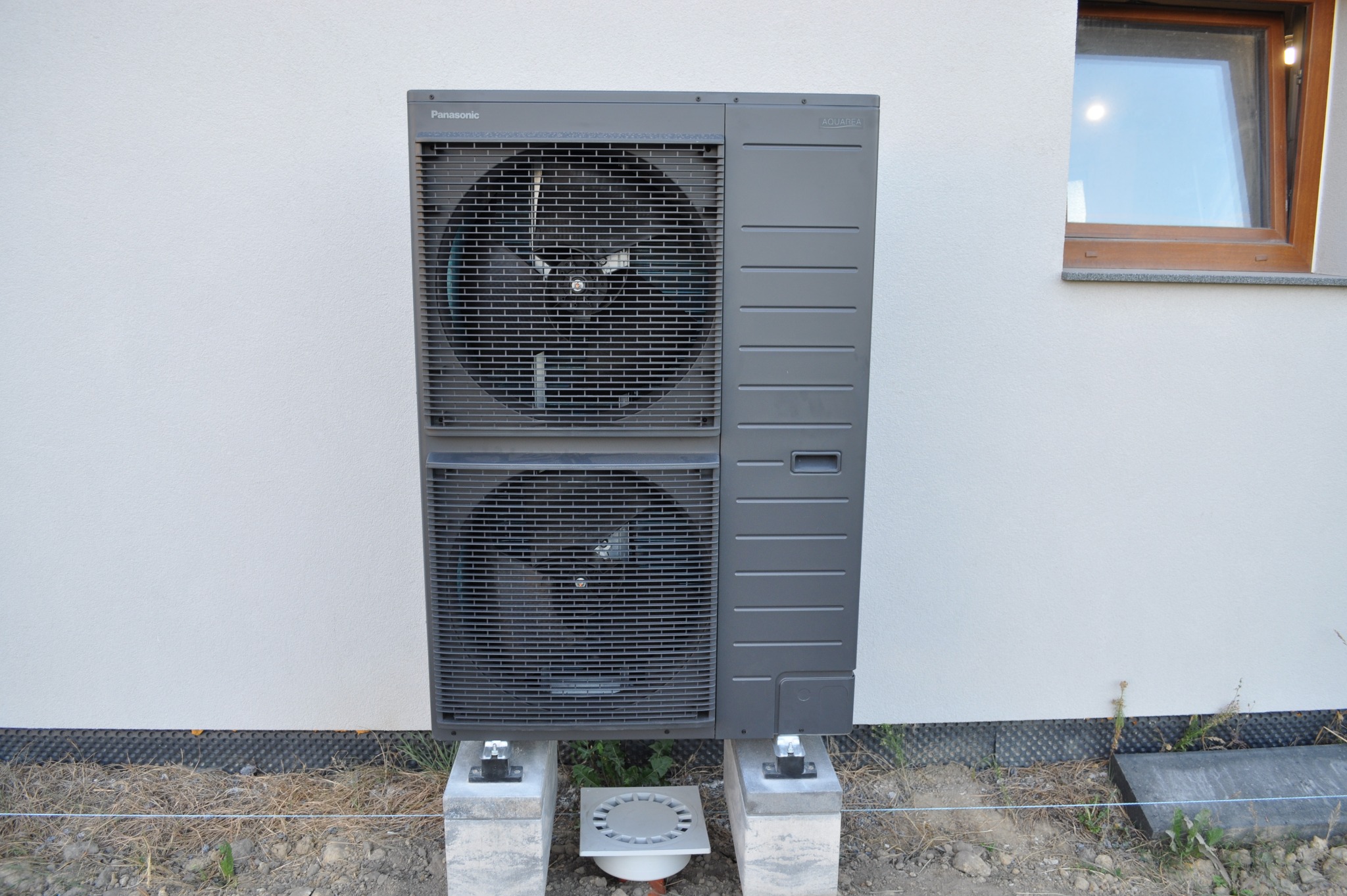 Szara pompa ciepła Panasonic Aquarea Split, zamontowana na betonowych blokach przy ścianie budynku z widocznym oknem. Widoczne elementy instalacji odprowadzającej skropliny.