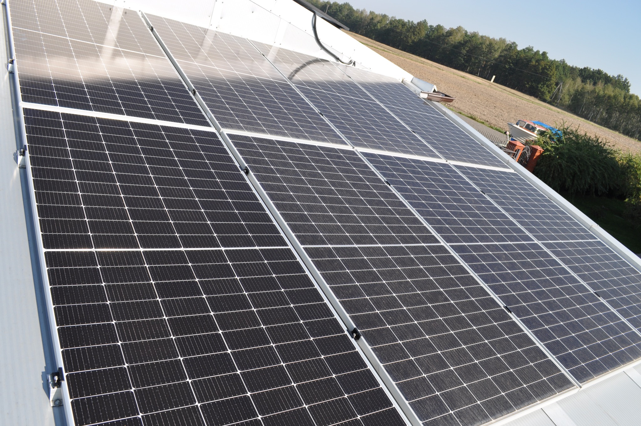 Panel słoneczny na dachu budynku w słoneczny dzień. Widoczne moduły fotowoltaiczne z bliska, z polami uprawnymi i drzewami w tle.