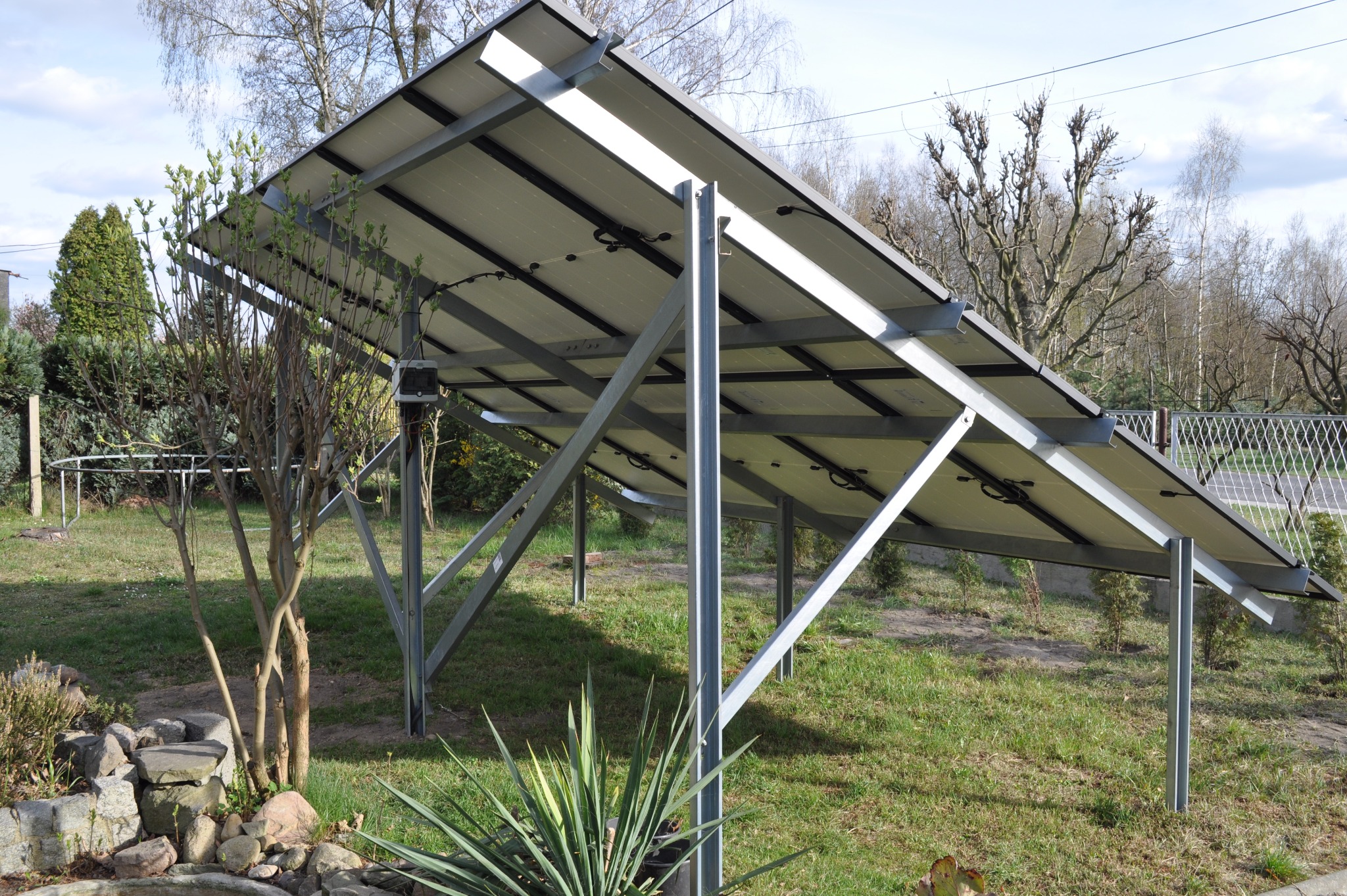 Zewnętrzny montaż paneli słonecznych na metalowej konstrukcji, widok od spodu, z zielenią w tle. Instalacja fotowoltaiczna na trawniku.