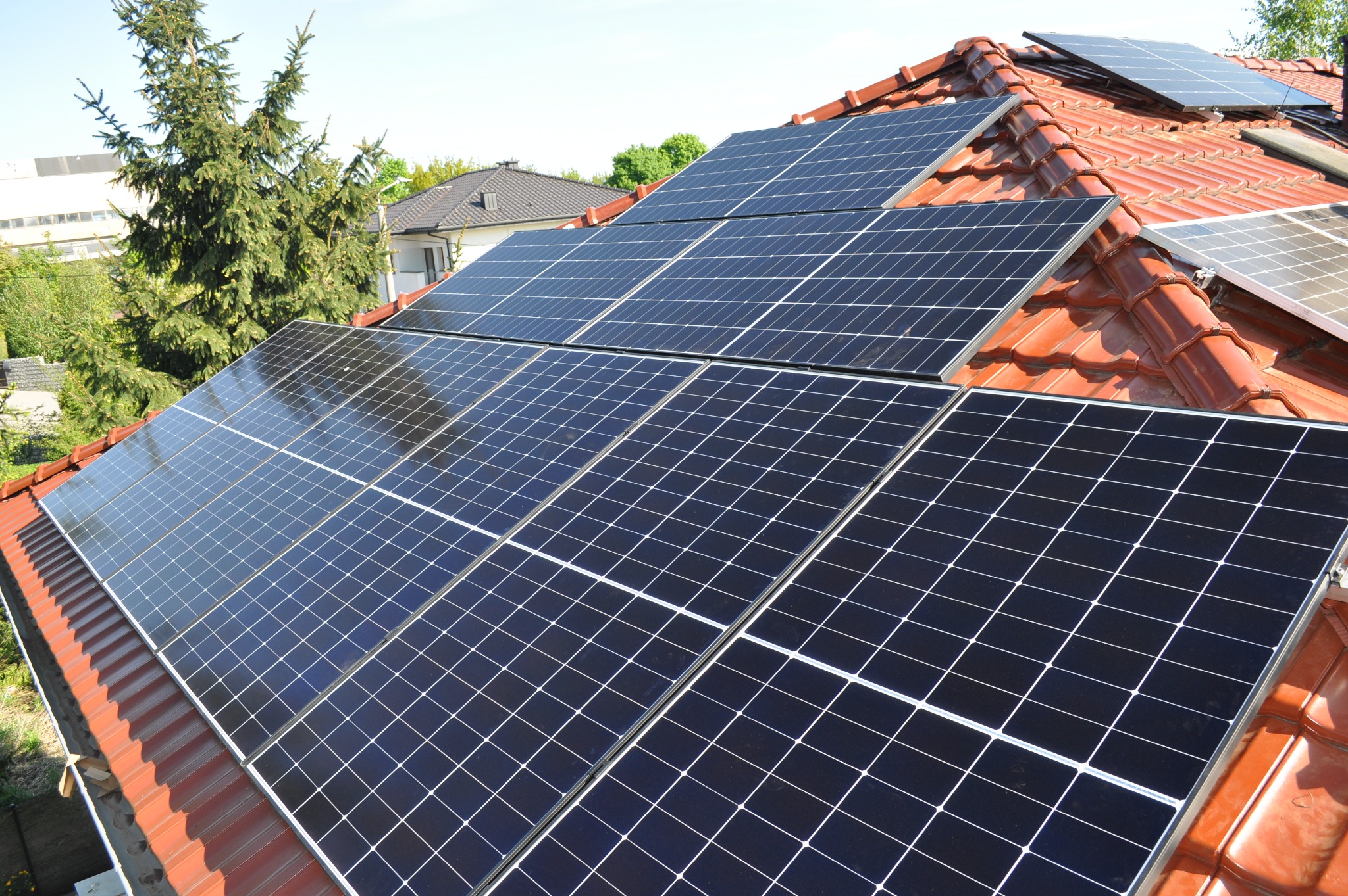 Instalacja fotowoltaiczna na dachu z czerwonej dachówki, widok z góry. Panele w układzie rzędowym, w tle zieleń drzew i budynki. Słoneczny dzień, błękitne niebo.