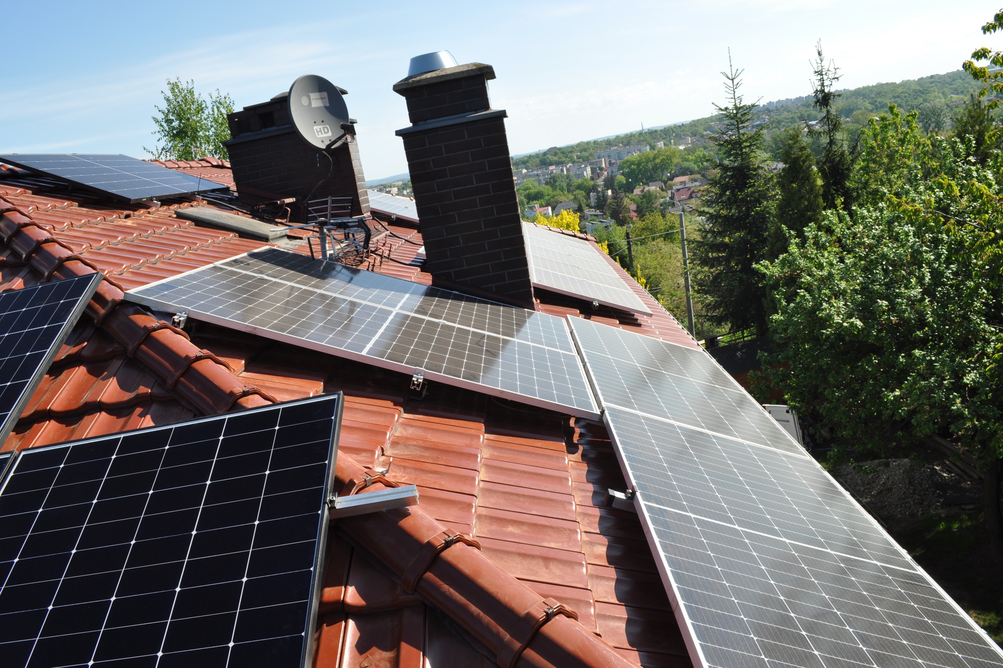 Panele fotowoltaiczne na dachu z cegły, komin i antena satelitarna w tle. Widok na zieleń i zabudowania miejskie w oddali, błękitne niebo.