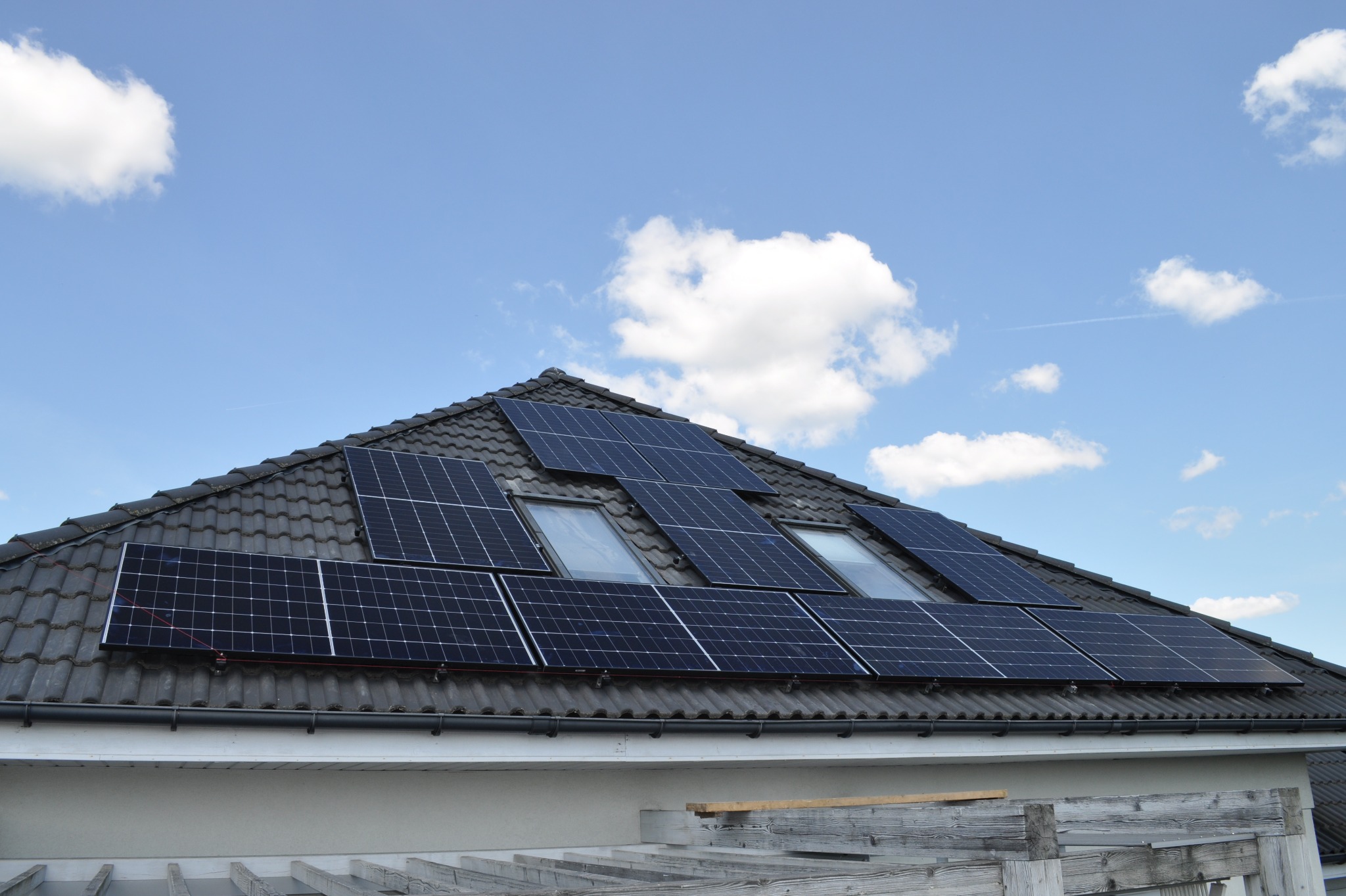 Panele słoneczne na dachu domu z dachówką, widoczne okna dachowe i fragment pergoli. Błękitne niebo z chmurami w tle. Instalacja fotowoltaiczna.