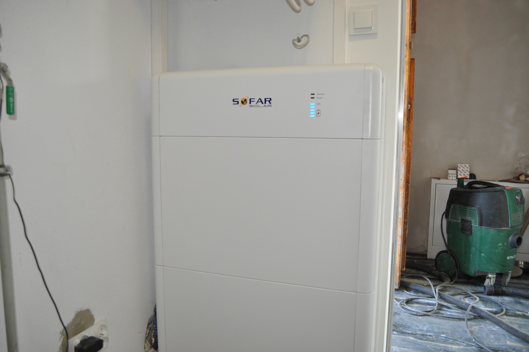 Biały akumulator energii SoFar Solar z diodami LED, widoczny w pomieszczeniu w trakcie instalacji. W tle odkurzacz przemysłowy i elementy wykończeniowe.