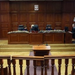 KANCELARIA ADWOKACKA ADW. ANDRZEJ KALISKI - Sala sądowa z sędziami w togach za stołem sędziowskim, publiczność na widowni. W tle drewniane panele i godło Polski.