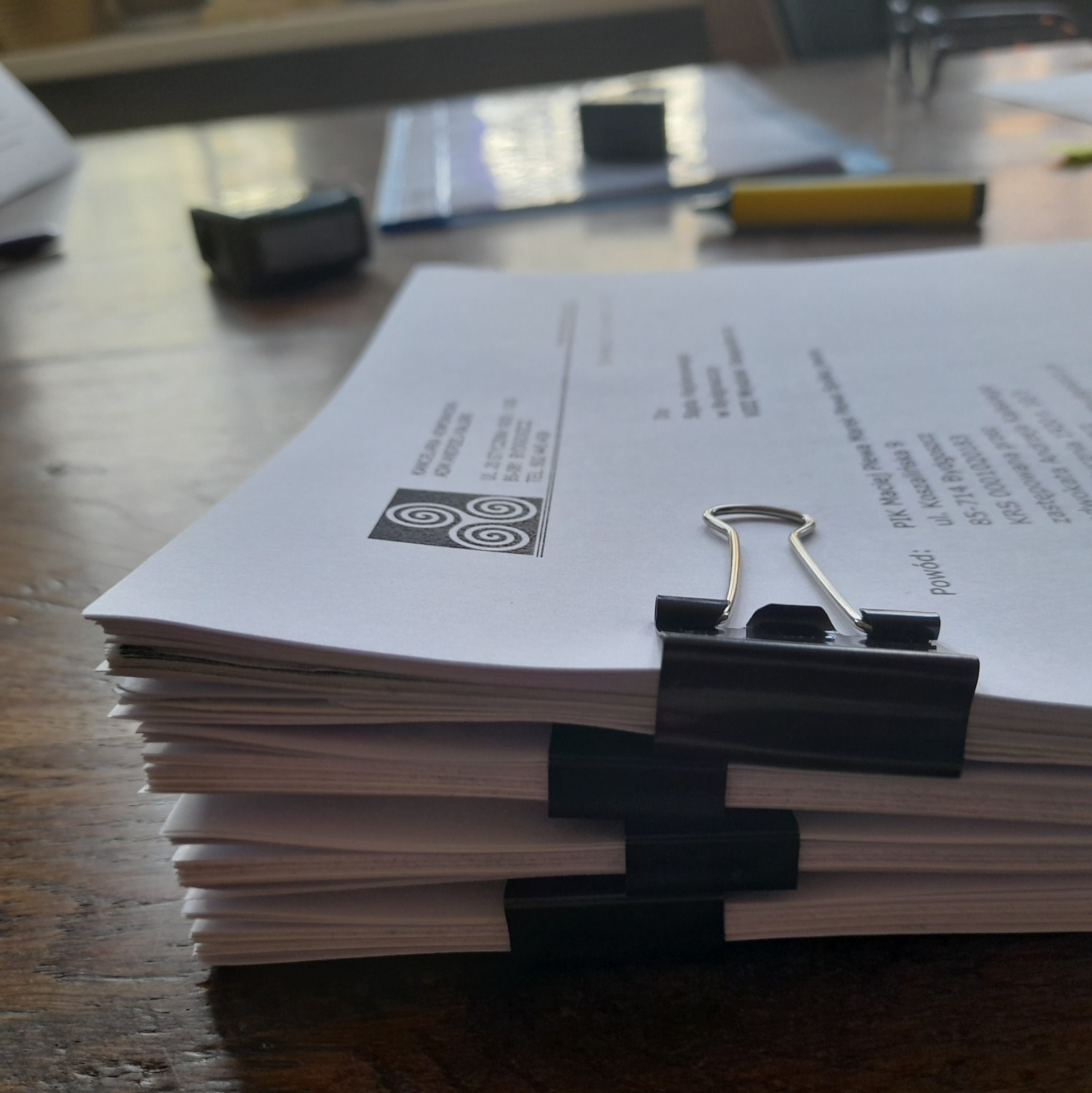 Stos dokumentów prawnych spięty czarnymi klipsami na drewnianym biurku, z widocznym logo i danymi Kancelarii Prawnej. W tle biurowe akcesoria.