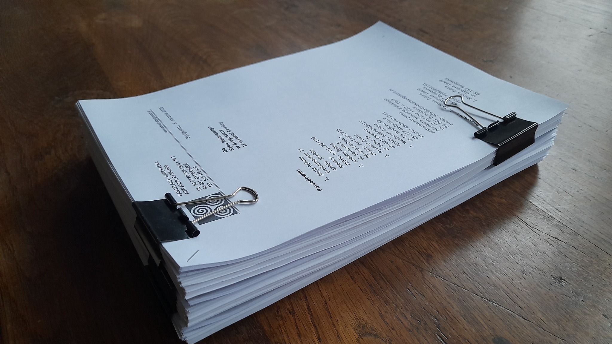 Stos dokumentów prawnych zabezpieczony dwoma klipsami biurowymi na drewnianym stole. Widoczne dane adresowe kancelarii adwokackiej i powodów.