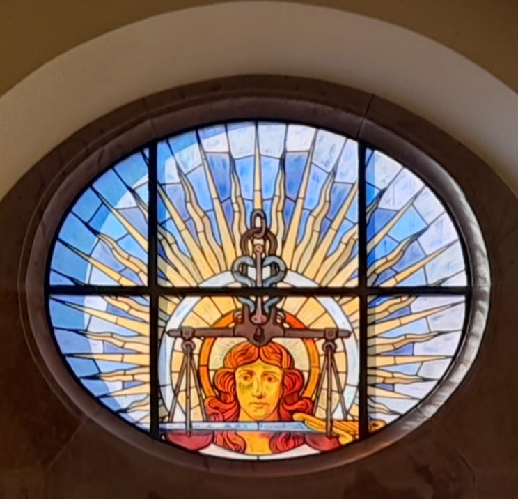Okrągły witraż w oknie prezentujący personifikację sprawiedliwości z wagą, otoczony promieniami na niebieskim tle. Symbol sądownictwa w artystycznym ujęciu.