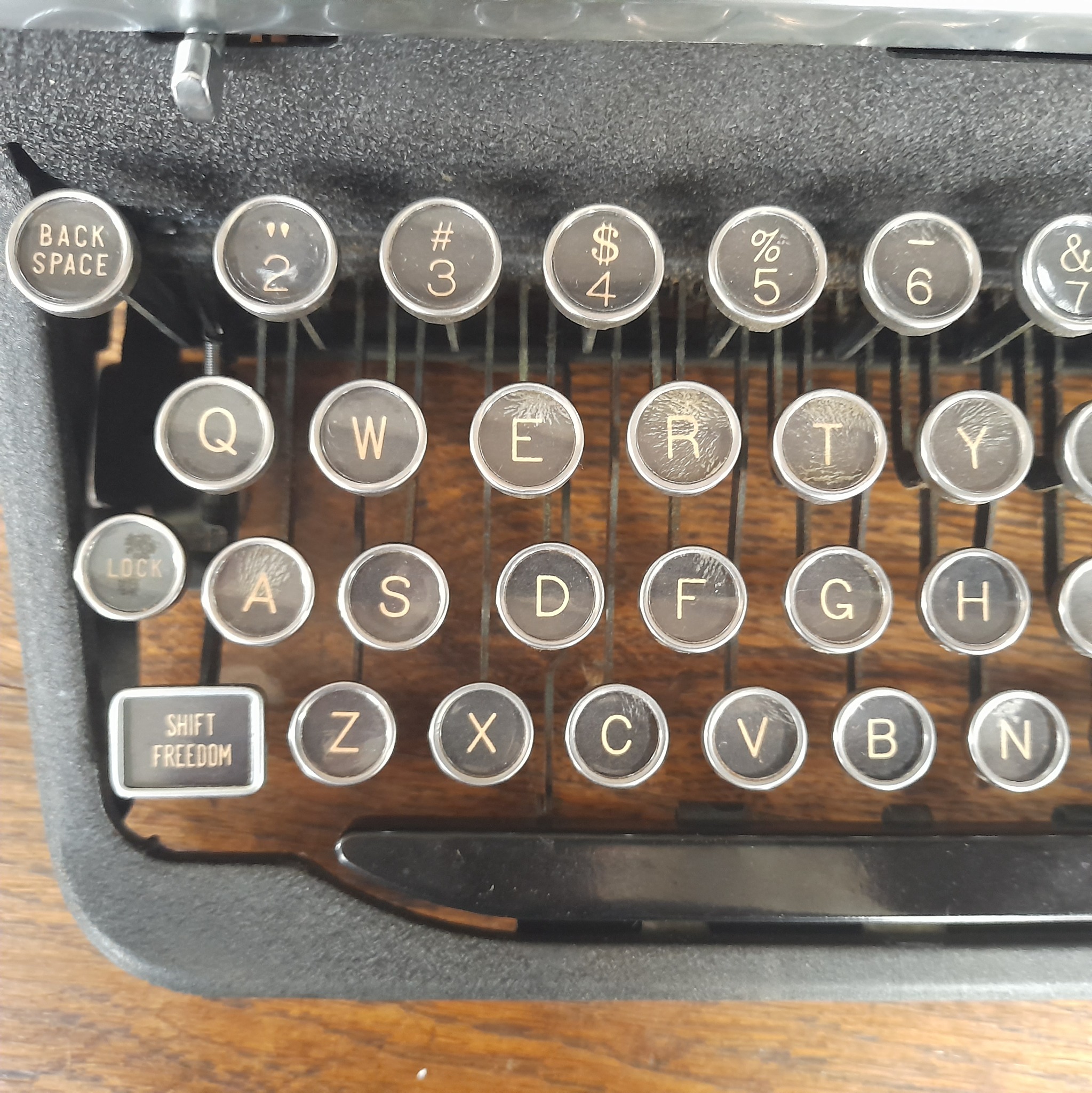 Czarno-srebrna, zabytkowa maszyna do pisania z okrągłymi klawiszami na drewnianym tle. Widoczne litery i cyfry na klawiszach.