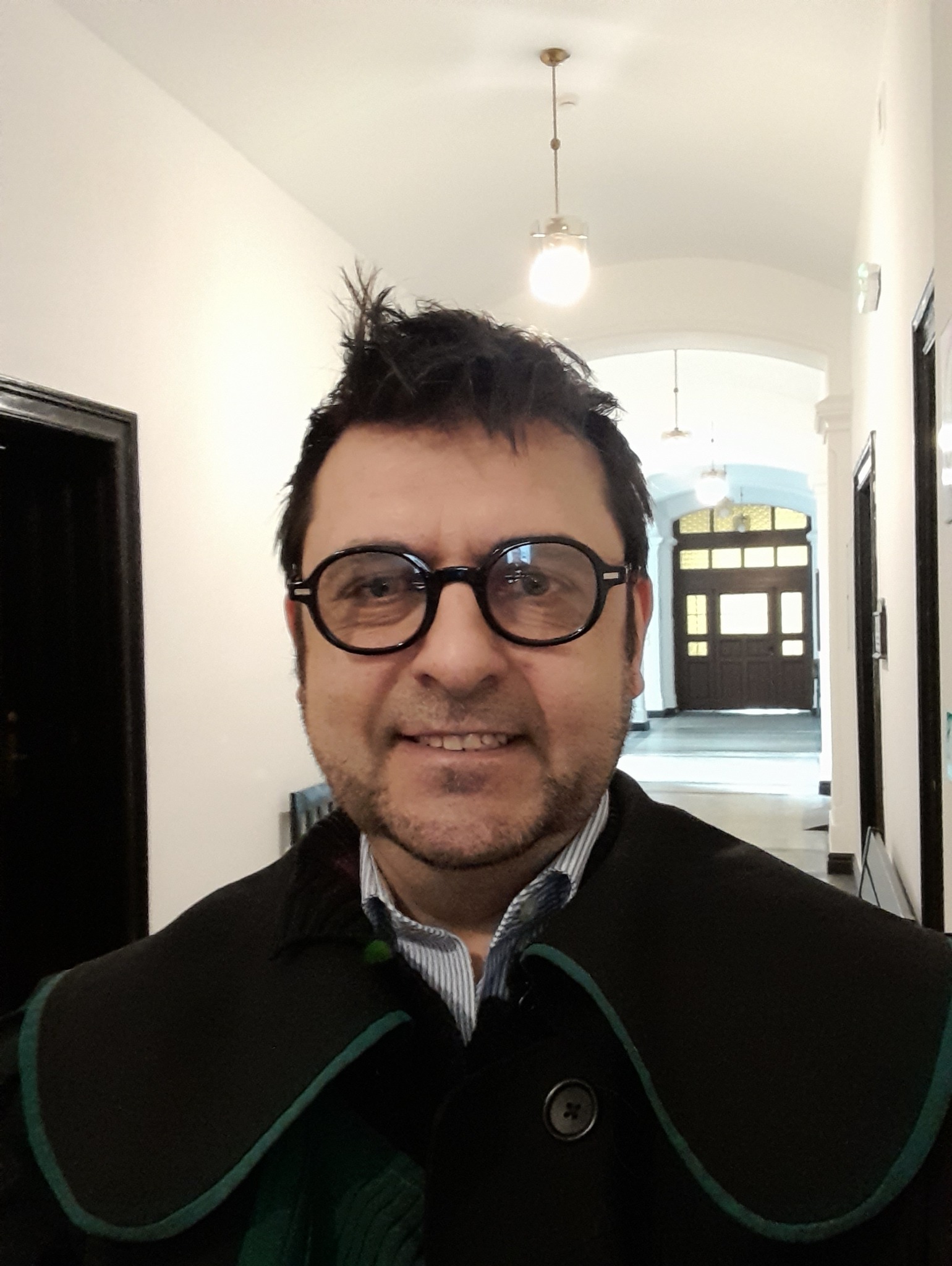 Portret uśmiechniętego mężczyzny w todze adwokackiej, w okularach, na tle korytarza z oświetleniem i drzwiami.