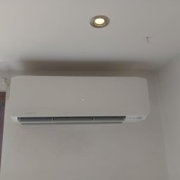 KlimaTech - Biała klimatyzacja Mitsubishi zamontowana na ścianie w jasnym pomieszczeniu, widoczna lampa sufitowa i fragment okna. Minimalistyczny design i schludny montaż.