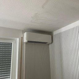 KlimaTech - Biały klimatyzator ścienny zamontowany w rogu pokoju z oknem i żaluzjami. Ściana w pionowe, szare pasy. Widoczne ślady na suficie.