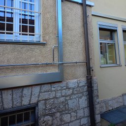 KlimaTech - Zewnętrzna instalacja odprowadzająca wodę deszczową, z metalowymi rurami i zabezpieczeniami okiennymi na fasadzie budynku z teksturą, widok z dołu.