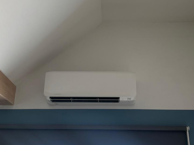 Biała klimatyzacja Mitsubishi zamontowana na ścianie w pokoju, widoczna część okna. Skośny sufit i drewniany element architektoniczny.