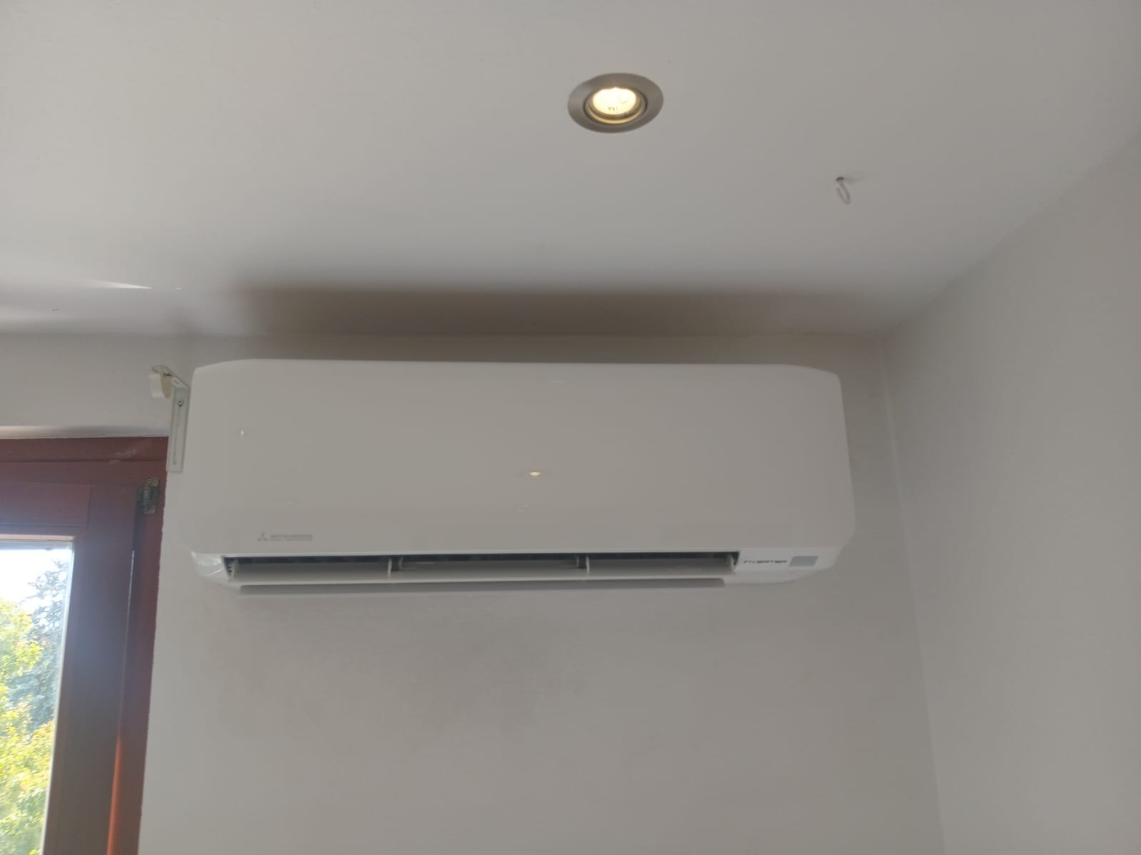 Biała klimatyzacja Mitsubishi zamontowana na ścianie w jasnym pomieszczeniu, widoczna lampa sufitowa i fragment okna. Minimalistyczny design i schludny montaż.