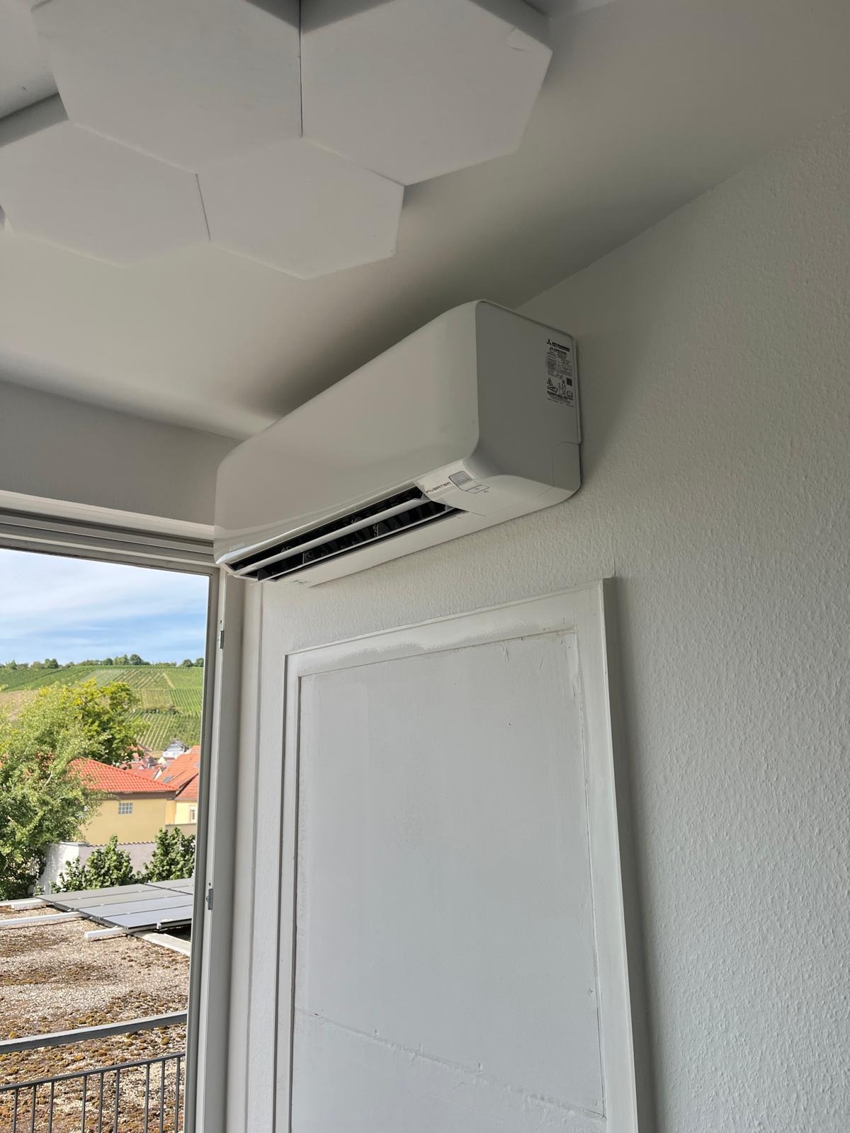 Nowoczesny klimatyzator Mitsubishi na ścianie w Karczmiskach Pierwszych, widok z okna na zielone wzgórza i zabudowania. Minimalistyczny design, białe wykończenie.