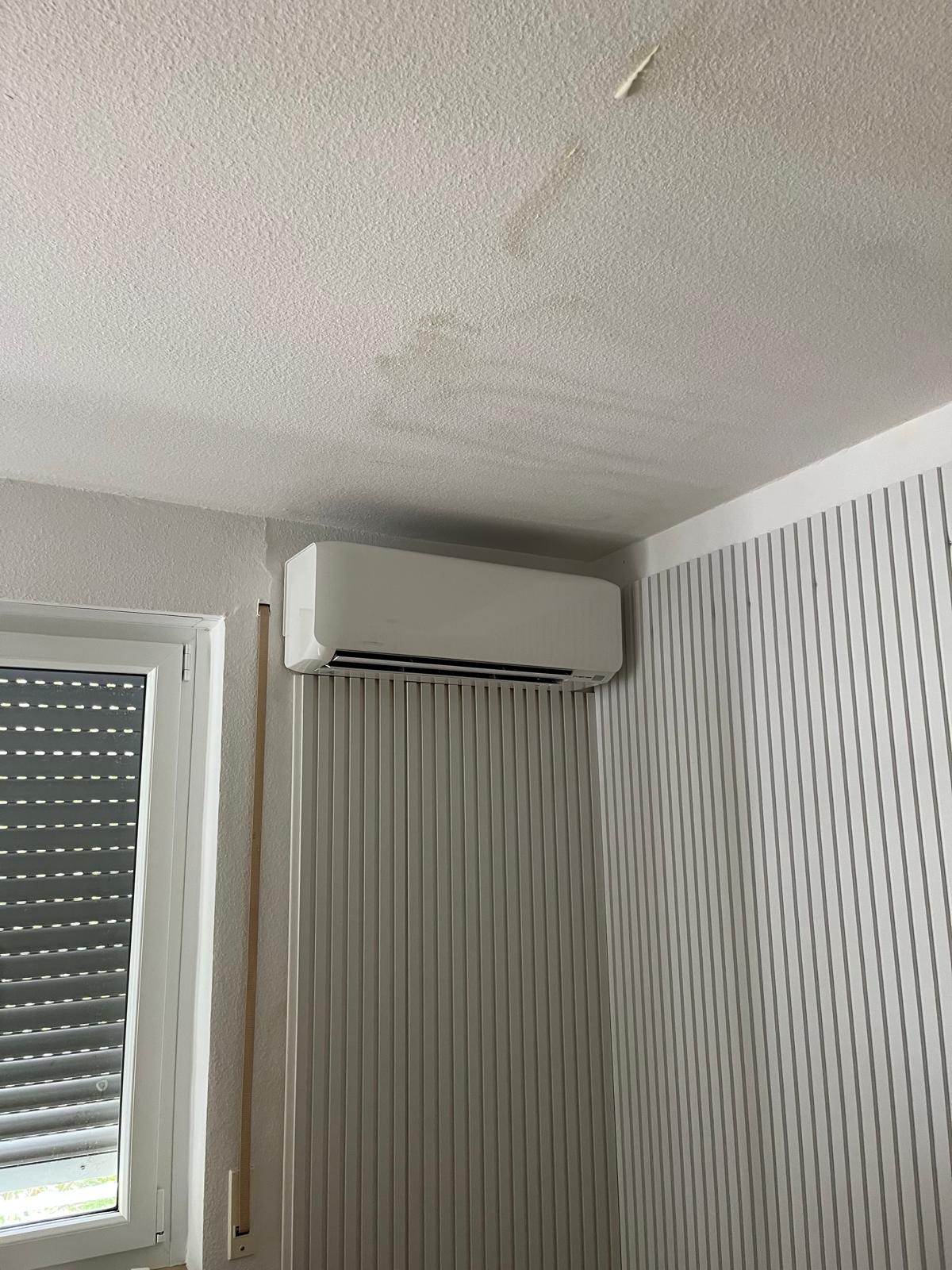 Biały klimatyzator ścienny zamontowany w rogu pokoju z oknem i żaluzjami. Ściana w pionowe, szare pasy. Widoczne ślady na suficie.