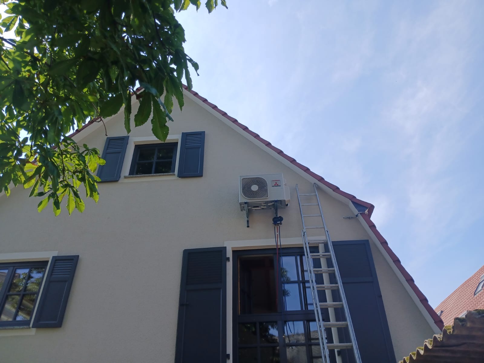 Biała jednostka zewnętrzna klimatyzacji Mitsubishi zamontowana na elewacji domu z szarymi okiennicami. Aluminiowa drabina oparta o ścianę. Widok z dołu na błękitne niebo.