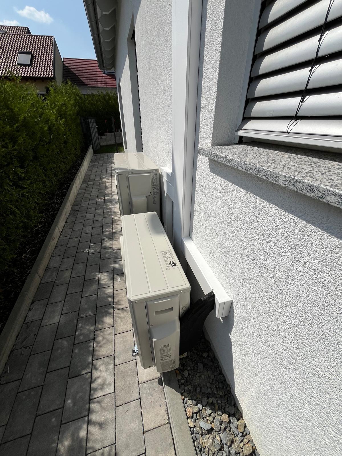 Dwa beżowe agregaty klimatyzacji zamontowane na zewnątrz budynku, widoczne z boku na tle białej ściany i kostki brukowej. Odpływ skroplin skierowany na kamienie.