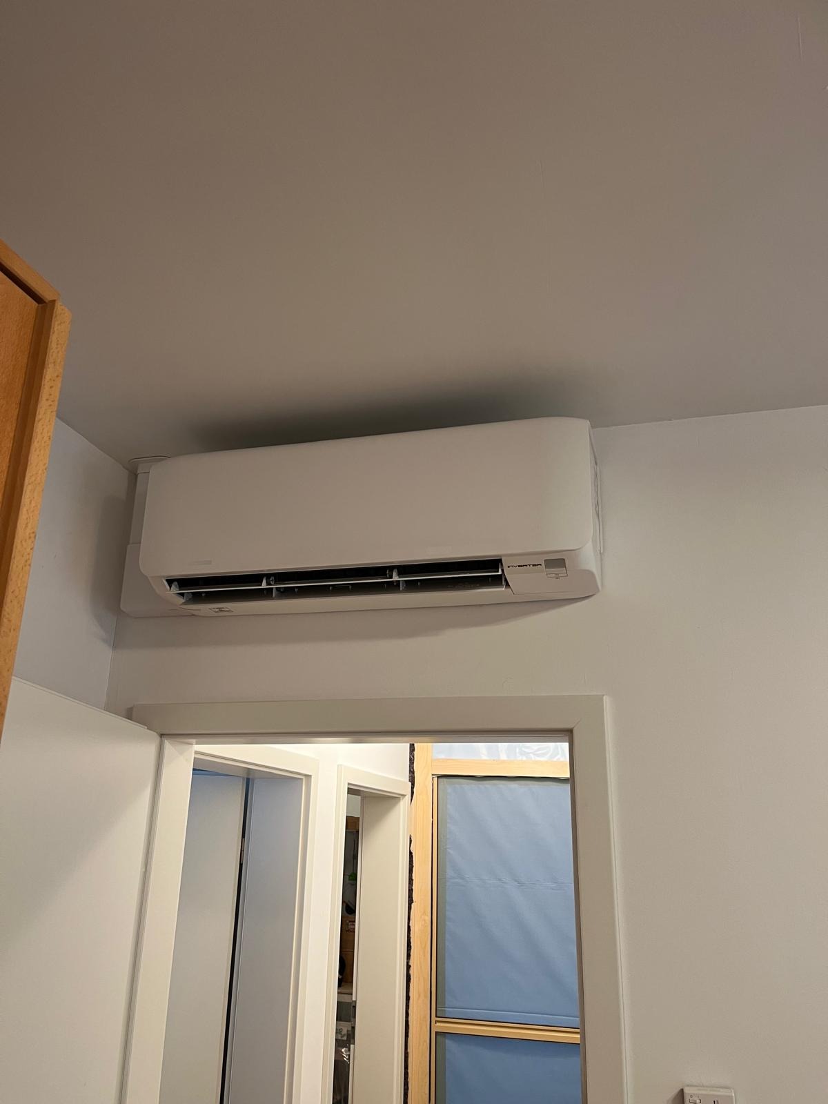 Biały klimatyzator typu split zamontowany nad drzwiami w jasnym pomieszczeniu. Widoczna instalacja. Minimalistyczny design.