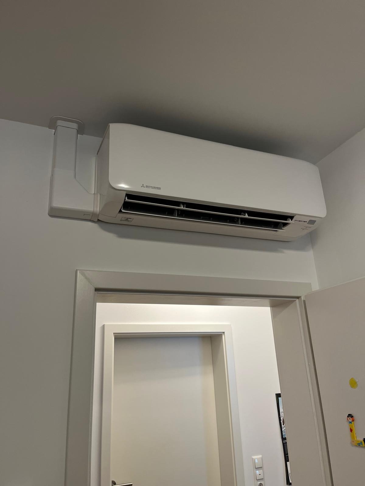 Biały klimatyzator Mitsubishi zamontowany nad drzwiami, widoczne elementy instalacji i białe ściany. Minimalistyczny design.