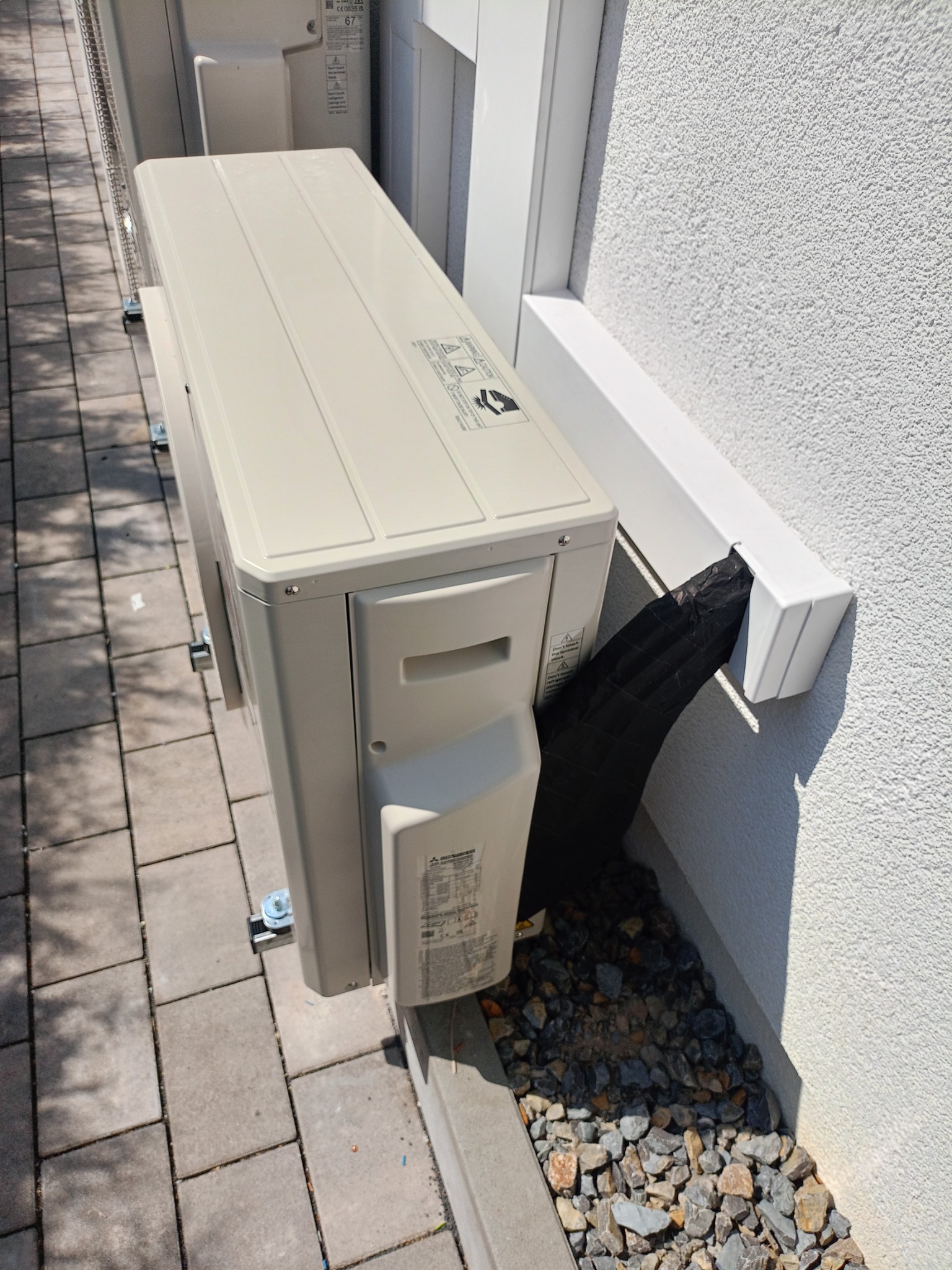 Zamontowana jednostka zewnętrzna klimatyzacji przy ścianie budynku z widocznym odprowadzeniem skroplin w osłonie. Widok z boku, w tle kostka brukowa i elewacja.