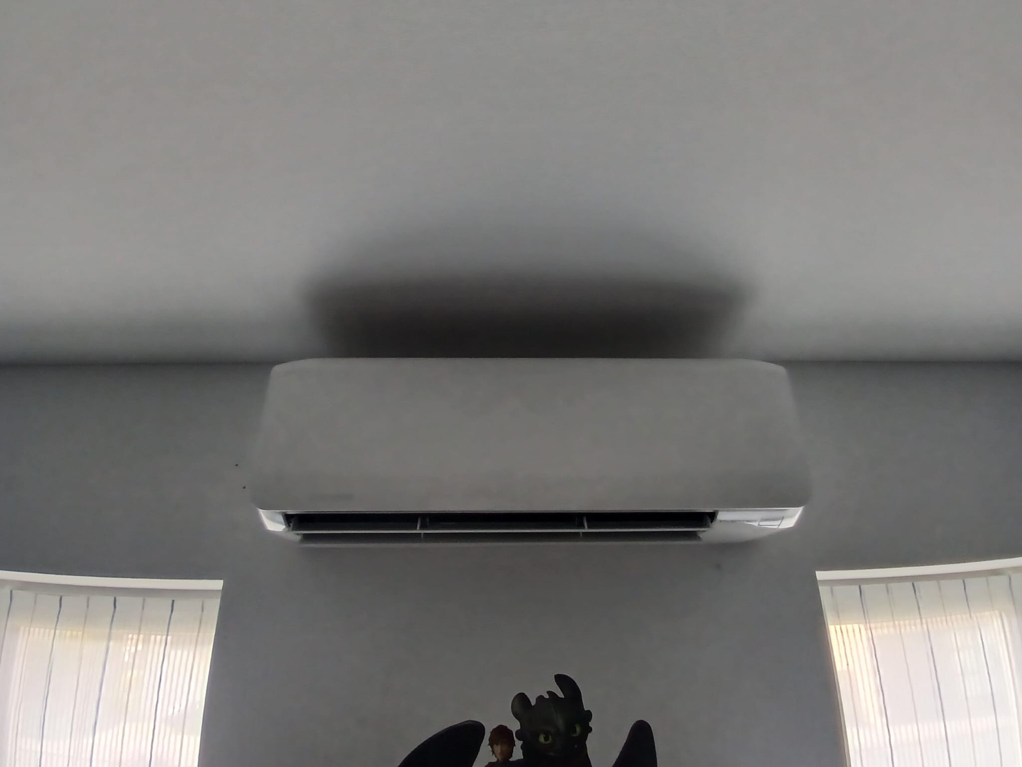 Biała klimatyzacja zamontowana na ścianie nad naklejką z motywem 'Jak wytresować smoka', widok z dołu. W tle fragment okna z żaluzjami.