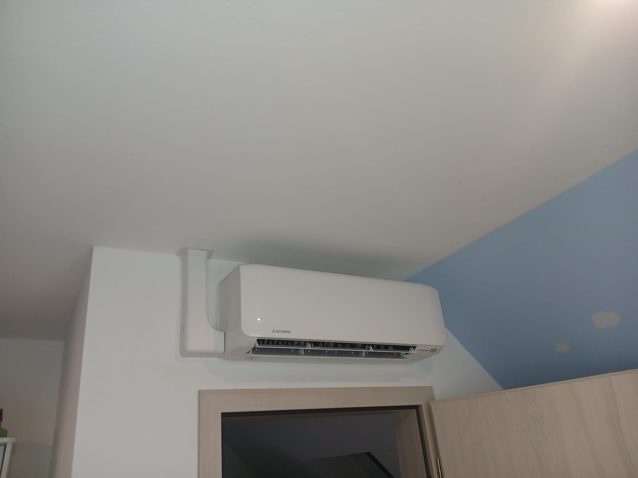 Biała klimatyzacja Mitsubishi zamontowana na ścianie nad drzwiami, z widoczną instalacją. Sufit biało-niebieski z motywem chmur. Widok z dołu.
