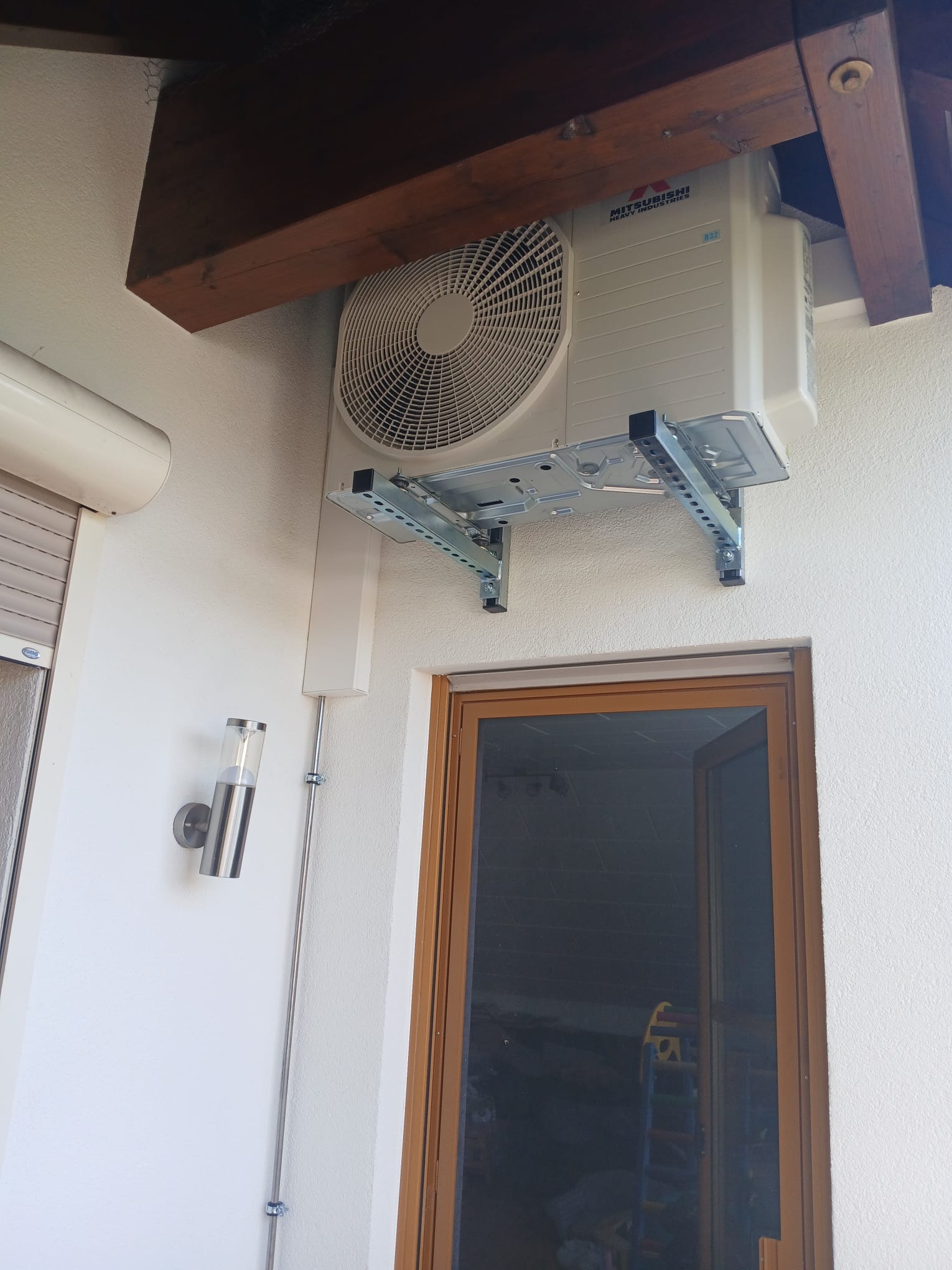 Biała jednostka zewnętrzna klimatyzacji Mitsubishi zamontowana na wspornikach pod drewnianym zadaszeniem, widok z dołu, drzwi balkonowe i lampa ścienna w tle.