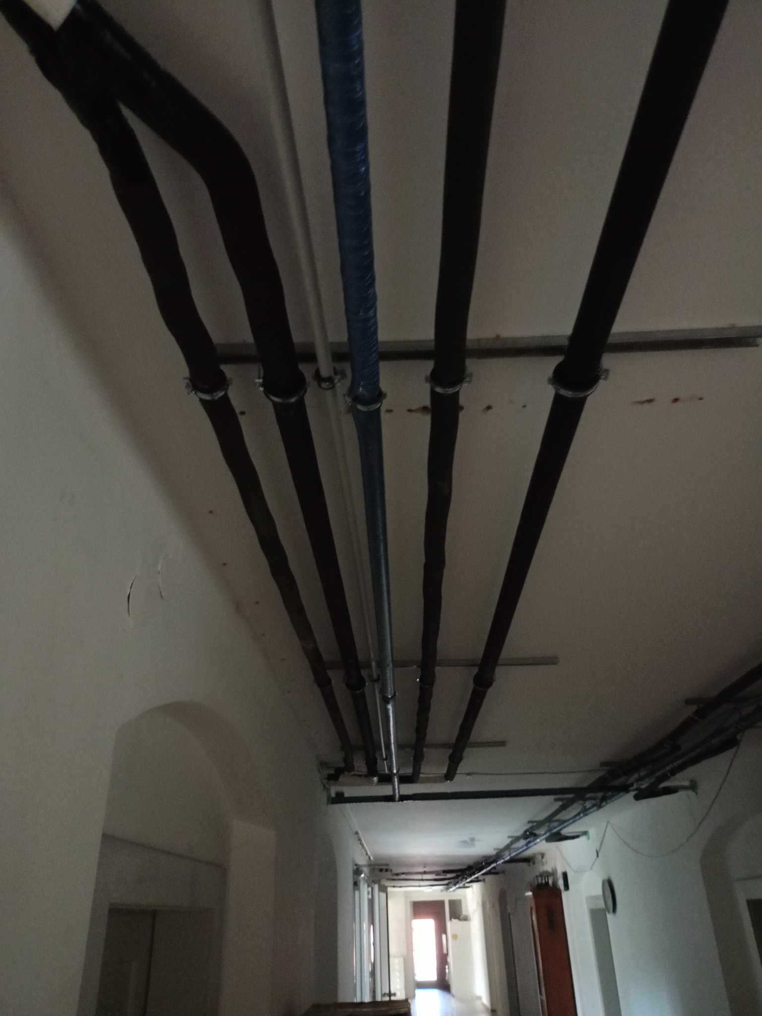 Instalacja rur pod sufitem w korytarzu, widoczne czarne i niebieskie rury, mocowania, fragment korytarza z łukowym przejściem.