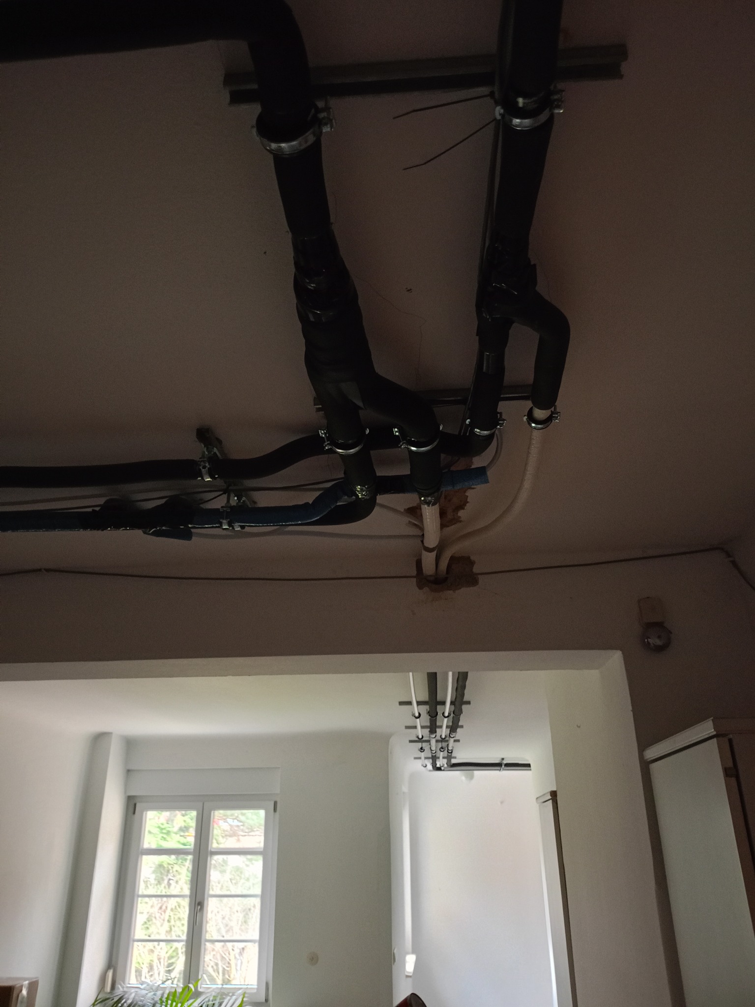 Czarna instalacja rur pod sufitem w Karczmiskach Pierwszych, mocowana metalowymi obejmami. Widok z dołu na pomieszczenie z oknem i rośliną.