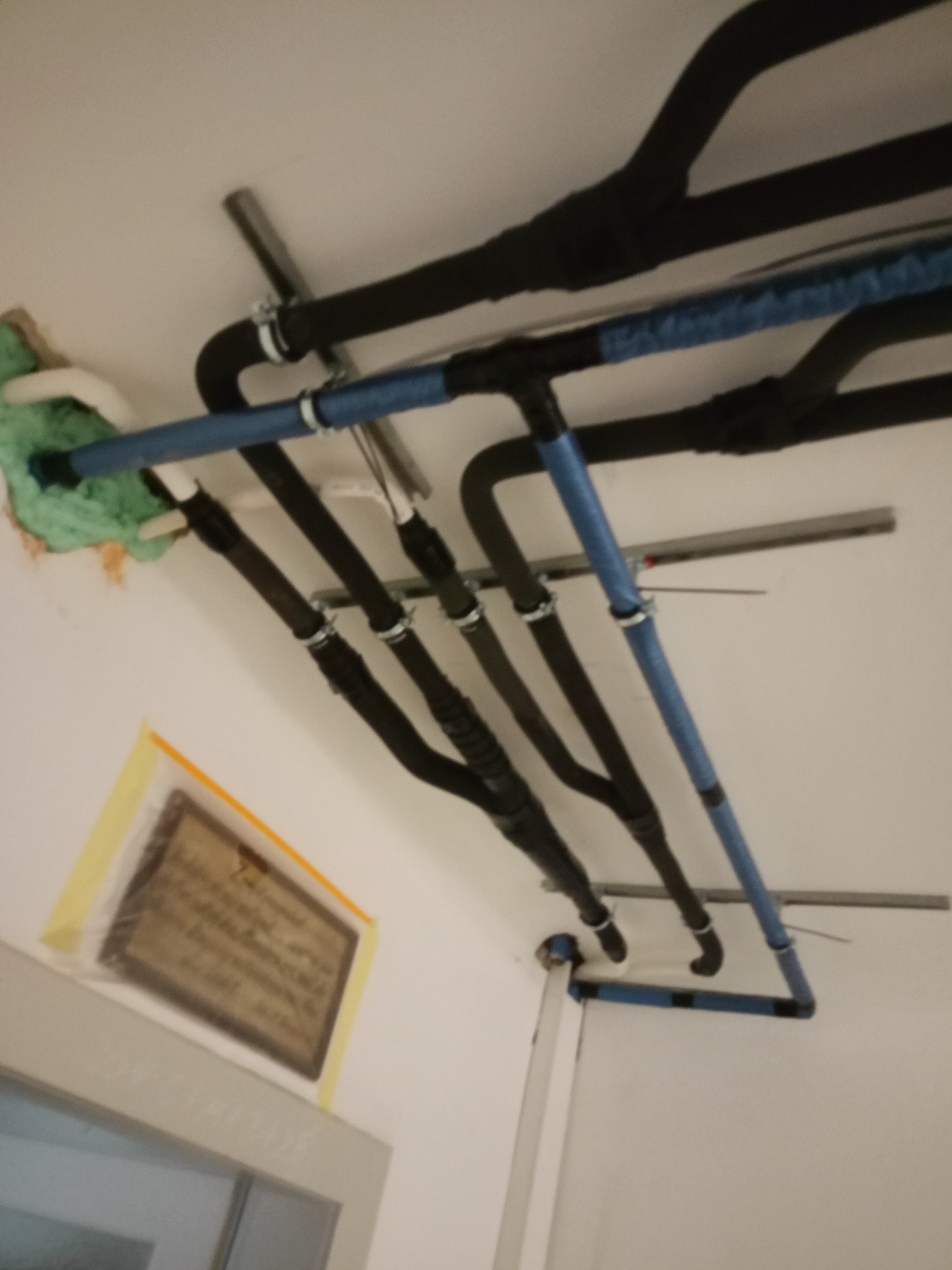 Instalacja rur w budynku, widoczne rury w izolacji termicznej, czarne i niebieskie, przymocowane do sufitu. Fragment ściany z oknem w dolnej części kadru.