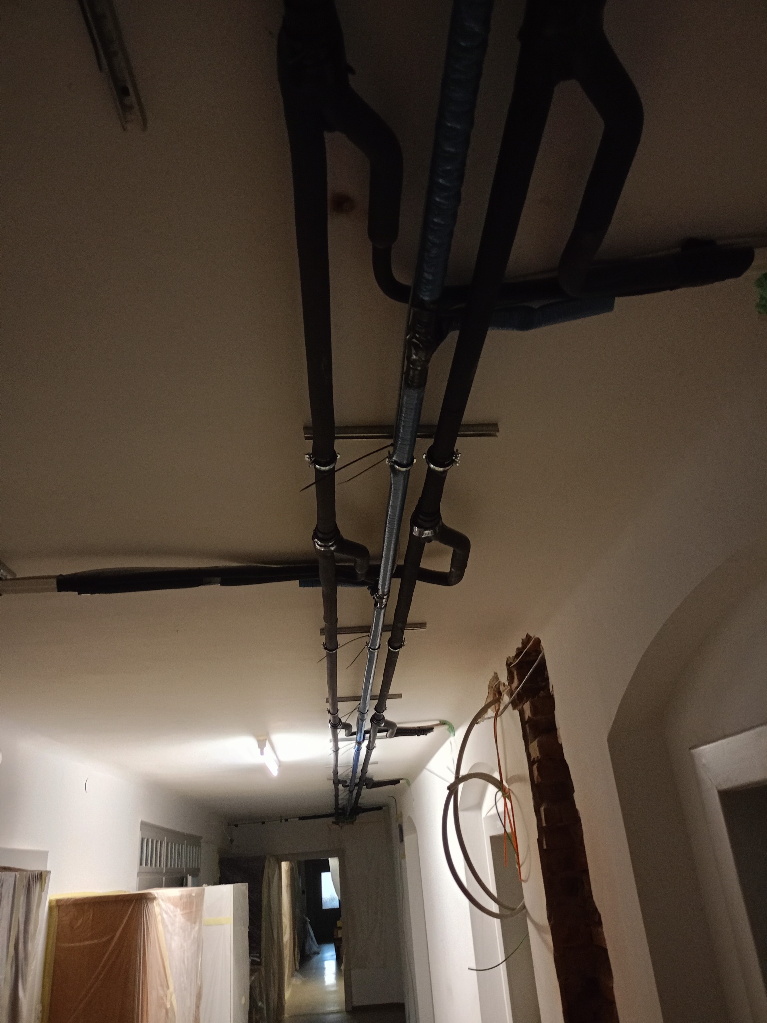 Instalacja rur wentylacyjnych pod sufitem w korytarzu, widoczne mocowania i izolacja termiczna. Częściowo wykończone wnętrze z elementami w trakcie prac.