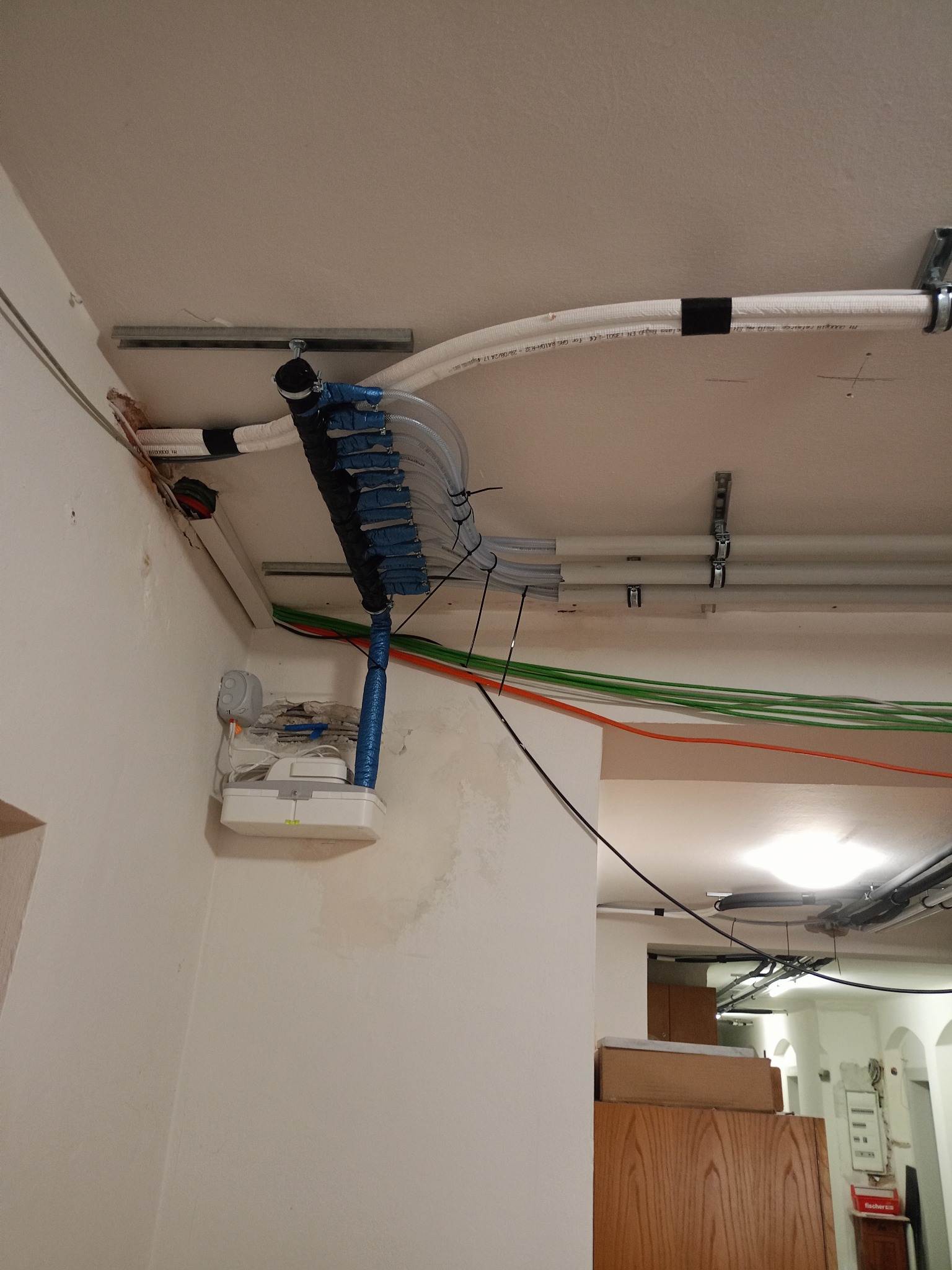 Instalacja rekuperacji pod sufitem z widocznymi rurami, przewodami i elementami montażowymi. Fragment ściany z puszką instalacyjną. Widok z dołu.