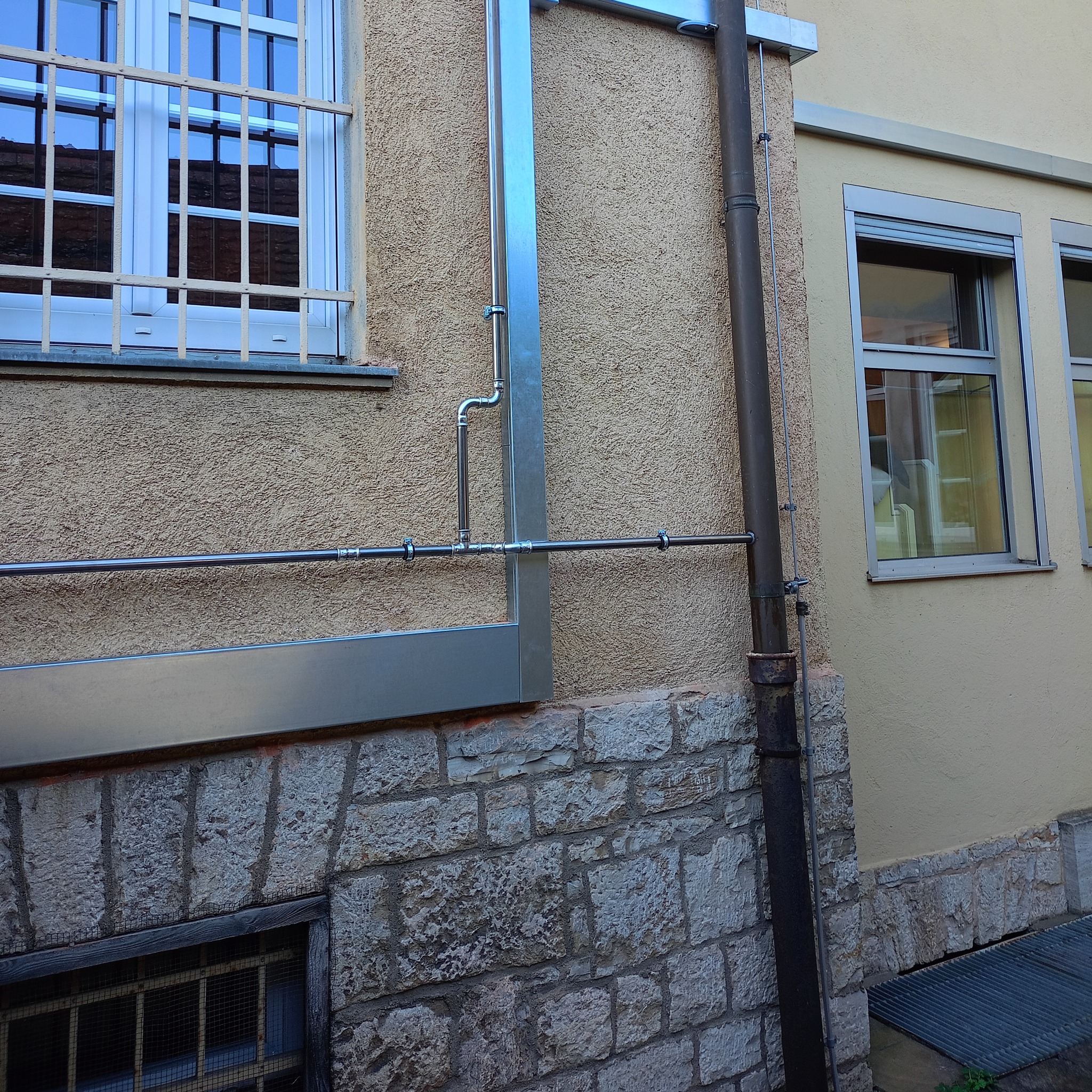 Zewnętrzna instalacja odprowadzająca wodę deszczową, z metalowymi rurami i zabezpieczeniami okiennymi na fasadzie budynku z teksturą, widok z dołu.