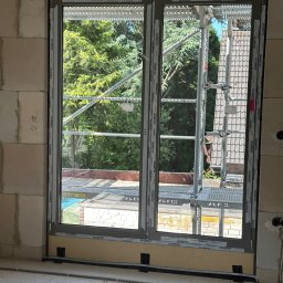 Marek Guga - Szare okno PCV w surowym murze z widokiem na zewnątrz, z widocznym rusztowaniem i zielenią. Okno zabezpieczone taśmą. W tle drzewa i dach budynku.