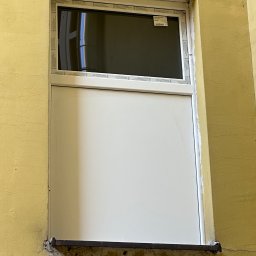 Marek Guga - Białe okno PCV z szybą u góry i panelem na dole, zamontowane w żółtej ścianie budynku. Widoczne ślady prac montażowych i starej instalacji.