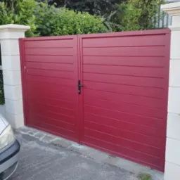 Czerwona brama wjazdowa z poziomymi panelami, widoczna z bliska, z fragmentem samochodu po lewej stronie. Tło stanowią krzewy i drzewa.