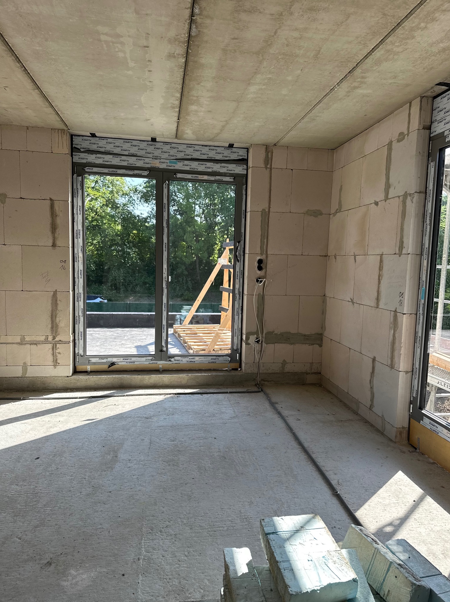 Surowy stan wnętrza z oknami PCV marki Aluprof, widoczne ślady po montażu, bloczki betonowe, instalacja elektryczna. W tle zieleń.
