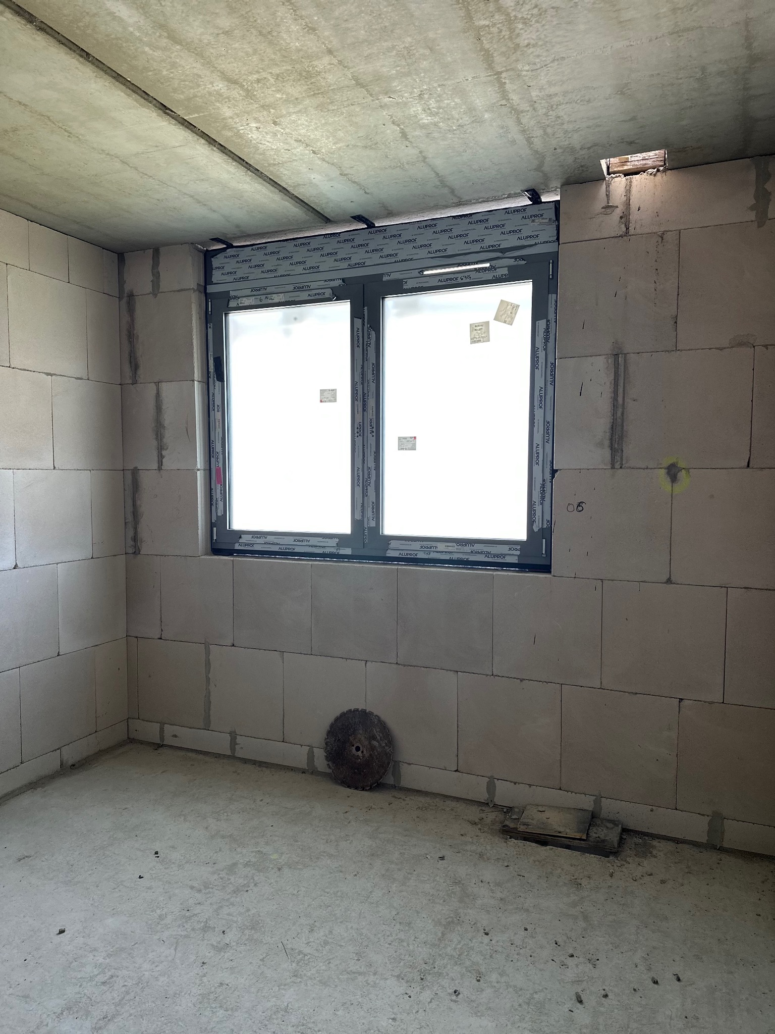 Szare okno z naklejkami w surowym, niewykończonym pomieszczeniu z bloków betonowych. Widoczne ślady po montażu.