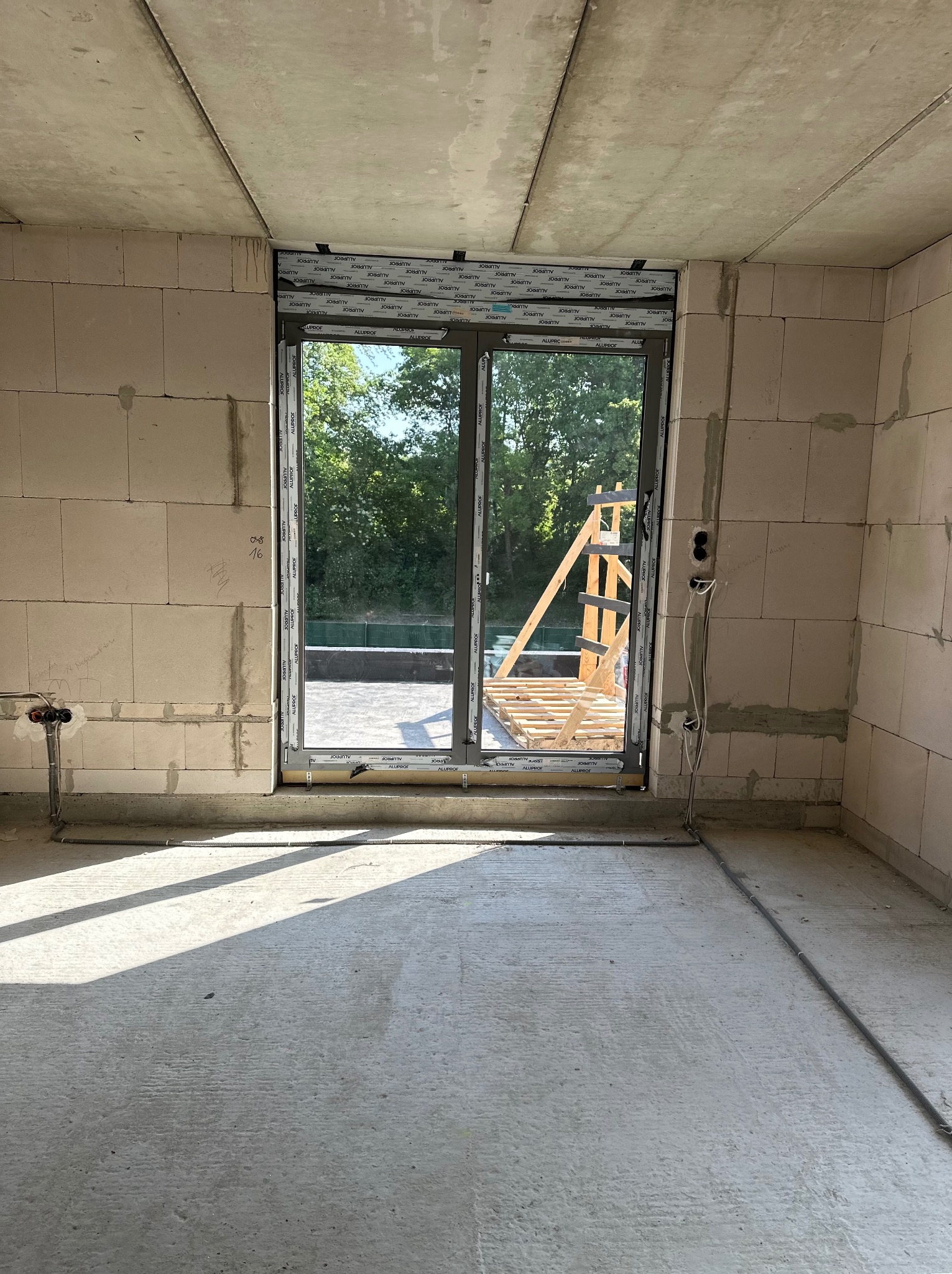 Surowy stan wnętrza z zamontowanymi oknami balkonowymi z widokiem na zewnątrz. Widoczne instalacje elektryczne i hydrauliczne, ściany z betonu komórkowego.