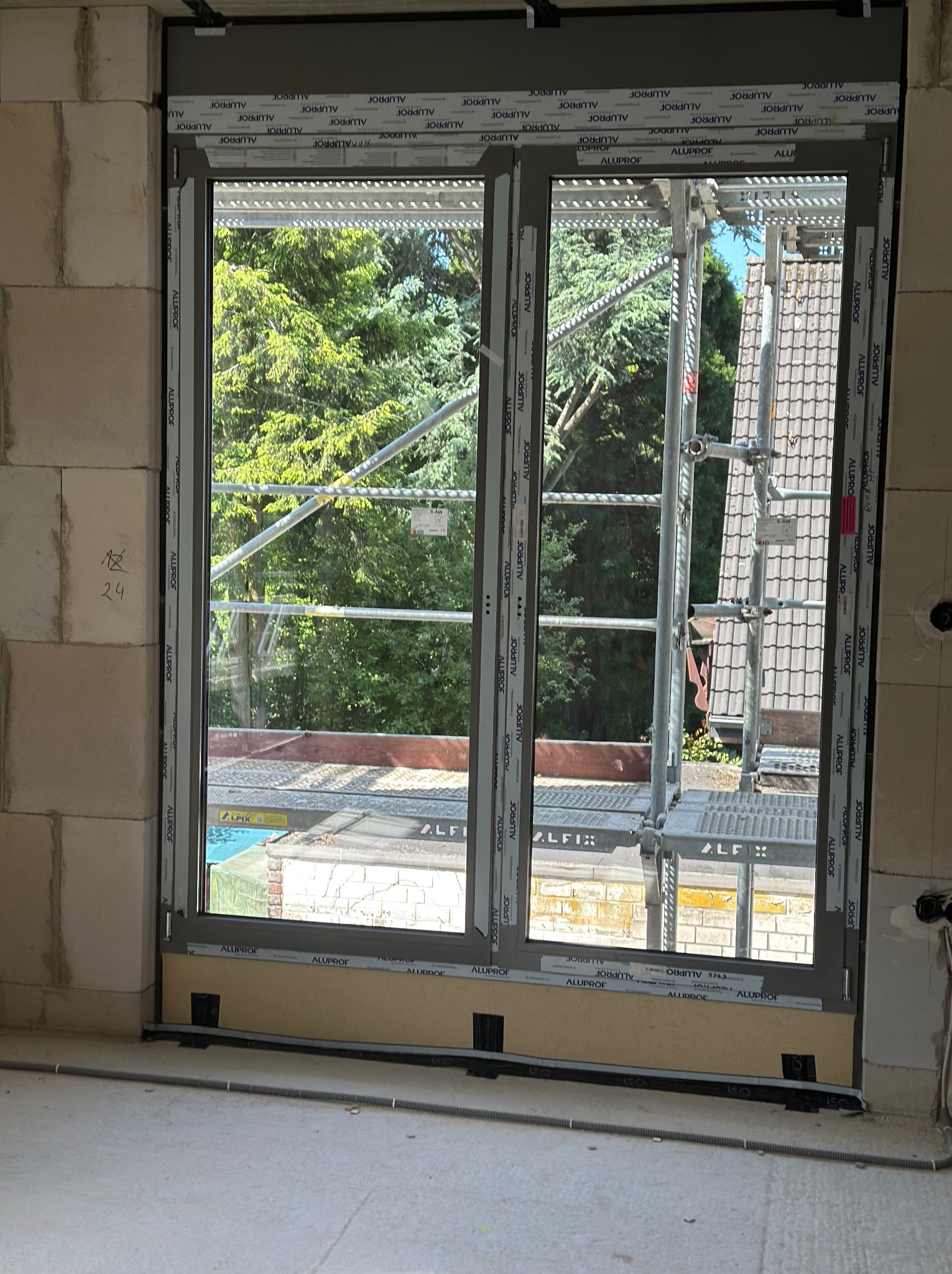 Szare okno PCV w surowym murze z widokiem na zewnątrz, z widocznym rusztowaniem i zielenią. Okno zabezpieczone taśmą. W tle drzewa i dach budynku.