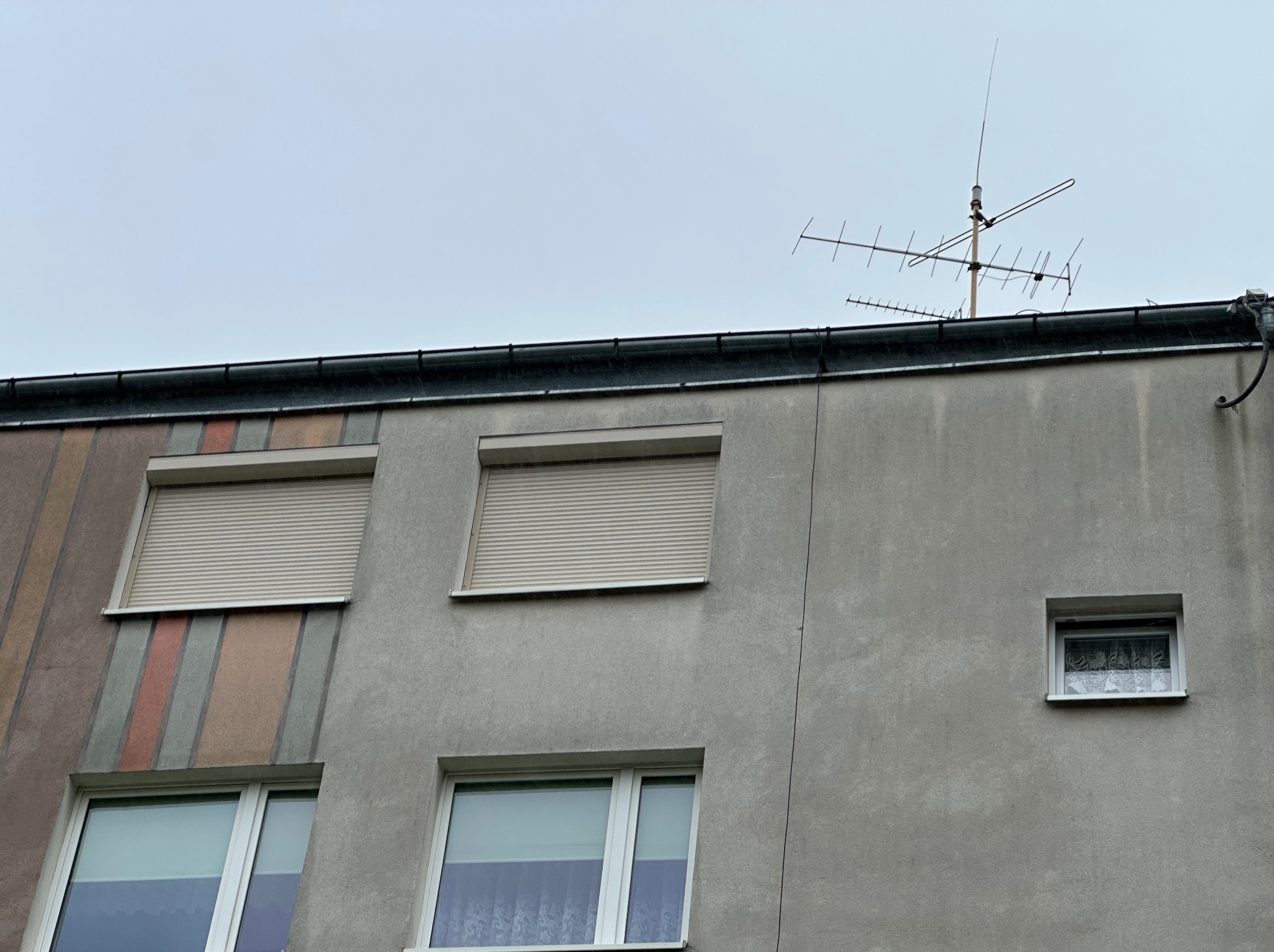 Fasada budynku z oknami i roletami zewnętrznymi w kolorze beżowym, z anteną na dachu. Widok na szary budynek w pochmurny dzień.