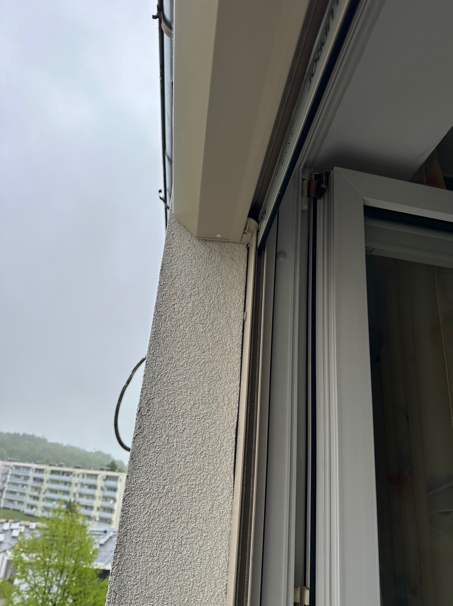 Fragment elewacji z zamontowaną prowadnicą rolety zewnętrznej, widok z dołu, zarys budynku w tle. Detal montażu.