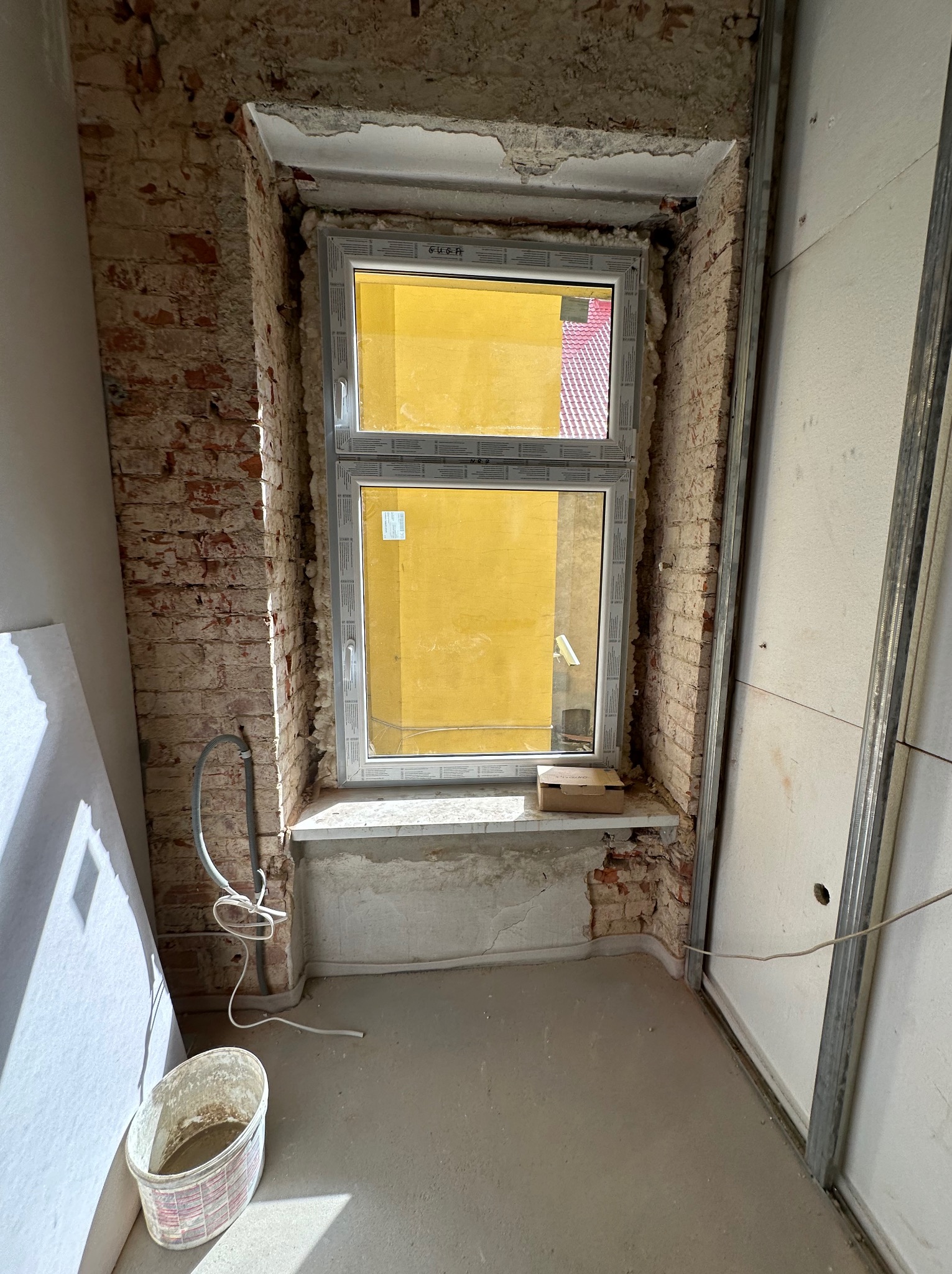 Montaż okna PCV w surowym wnętrzu. Widoczne cegły, instalacja elektryczna i gładzie. Prace wykończeniowe w toku, okno z widokiem na żółtą elewację.