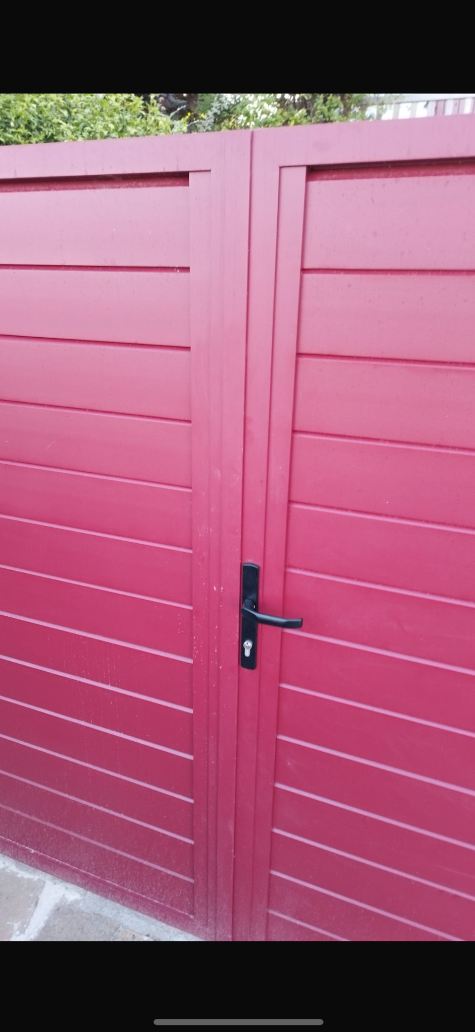 Czerwona, pozioma brama garażowa z czarną klamką. Widoczne poziome panele i pionowe wzmocnienia. Zewnętrzna brama w kolorze ceglastym.